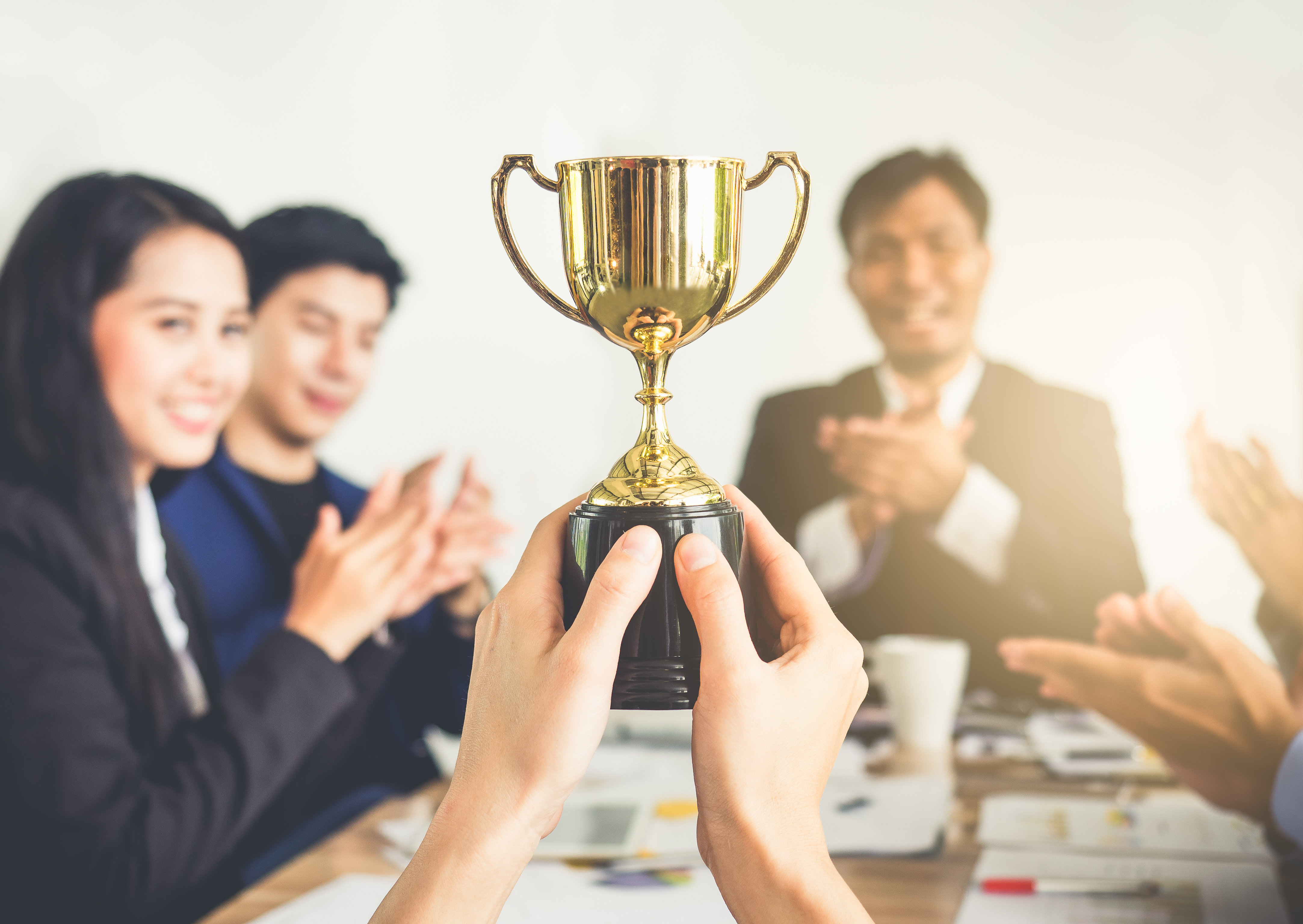 Person holding up a trophy and a group of employees clapping around a table looking at it