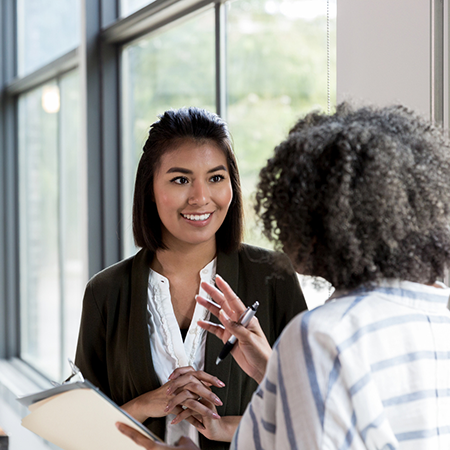 Two women talking while one is smiling and the other is holding a pen and notebook